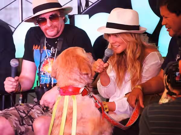 Comedian Rhys Darby looks over as Kristen Bell embraces the Dancing Merengue Dog during a press conference at Bonnaroo 2012.