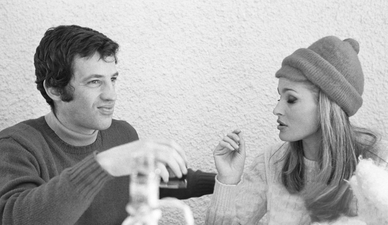 Jean-Paul Belmondo et Ursula Andress assis à table ensemble sur une terrasse en hiver, habillés chaudement. Il regarde loin devant lui et elle baisse les yeux,  (1967)