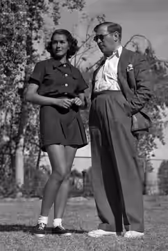 Jinx Falkenburg and Gregory Ratoff stand together in Palm Springs. She wears shorts, he wears pants, a jacket, and sunglasses. Both wear sneakers. Trees and a f