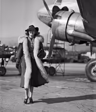 Actress Phyllis Brooks at an airfield wearing a fur-lined coat and a fedora, with an airplane visible in the background. Brooks was originally known as the "Ipa (1937)