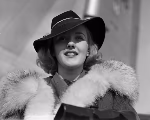 Actress Phyllis Brooks wearing a fur-lined coat and a fedora, with an airplane visible in the background. Brooks was originally known as the "Ipana Toothpaste G (1937)