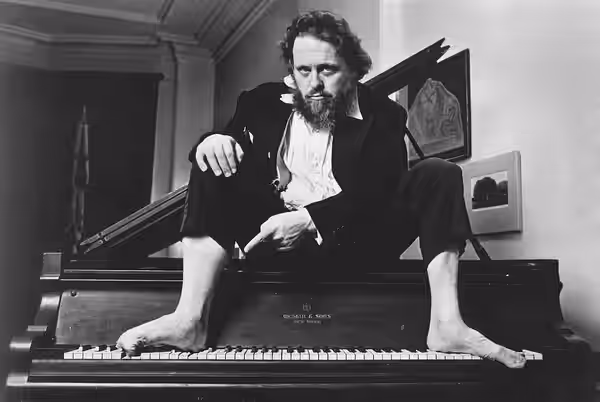 Peter Schickele seated barefoot atop a piano (1983)