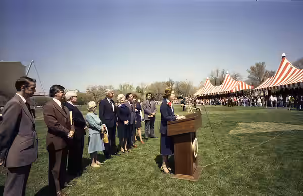 Scope and content: This photograph depicts First Lady Betty Ford standing at a lectern delivering remarks to a crowd seated under striped tents. The group stand