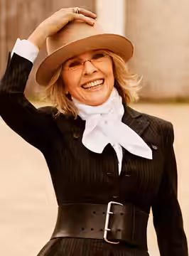 American actress Diane Keaton, smiling in a tan hat, white scarf, and black pinstriped jacket with a wide belt, her hand resting on her hat. (2012)