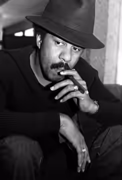 Portrait of American comedian Richard Pryor, seated, wearing a fedora, and holding a cigar (1976)