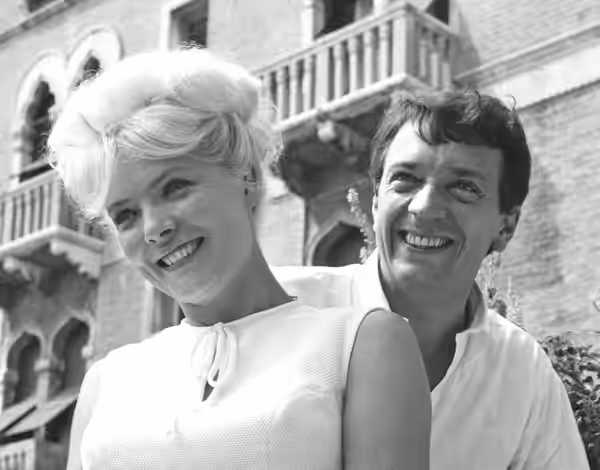 À l'extérieur devant un palais vénitien, Corinne Marchand devant Jean-Pierre Cassel, tous deux très souriants. Elle porte les cheveux blonds en chignon et est v (1962)