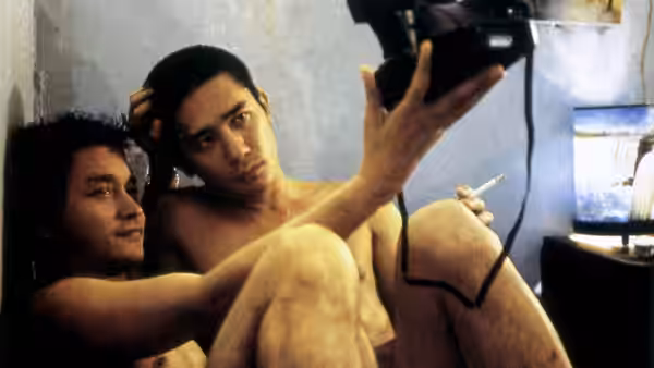 Two shirtless men sit on the floor holding a video camera