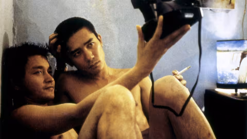 Two shirtless men sit on the floor holding a video camera
