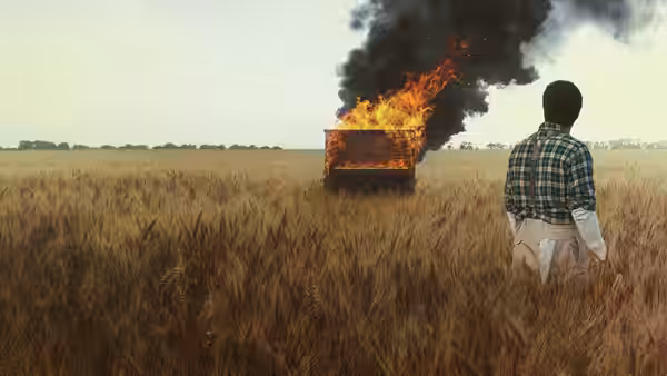 A man watches a burning vehicle from a wheat field