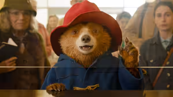 Paddington (Ben Whishaw) waving through glass in an airport terminal