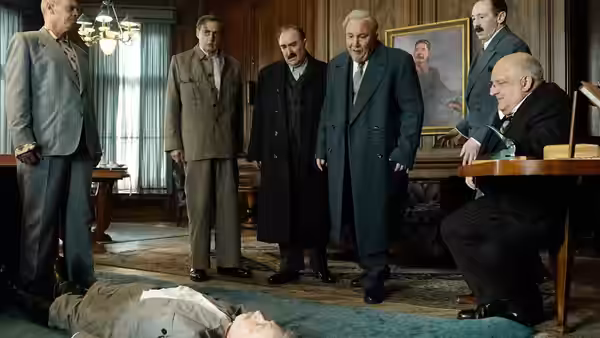 Men in suits and coats stand over a body on the office floor