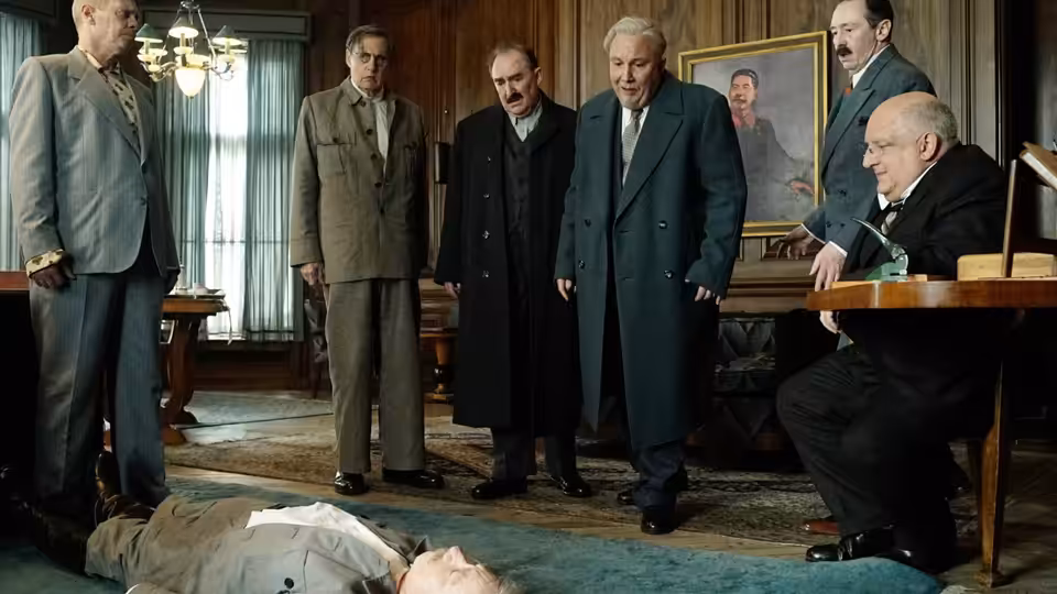 Men in suits and coats stand over a body on the office floor