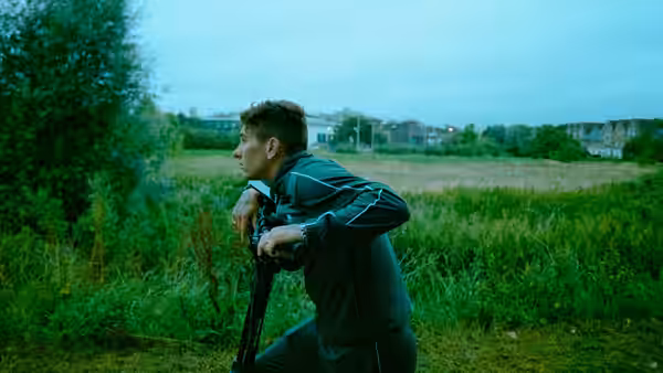 A man kneels in tall grass holding a scooter while looking sideways across a field