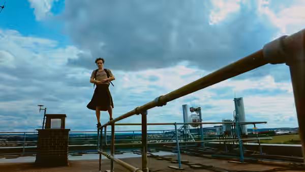 A woman balances on a metal railing on a rooftop overlooking a city under clouds