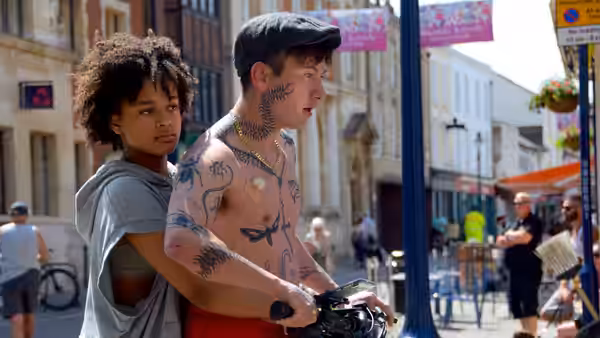 A young woman stands close behind a shirtless tattooed man on a bicycle in a street