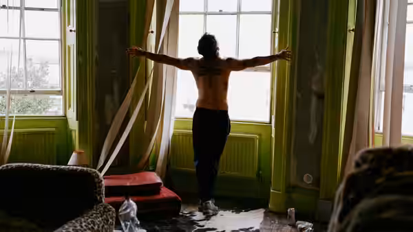 A shirtless man with back tattoos stands with arms outstretched in a derelict room