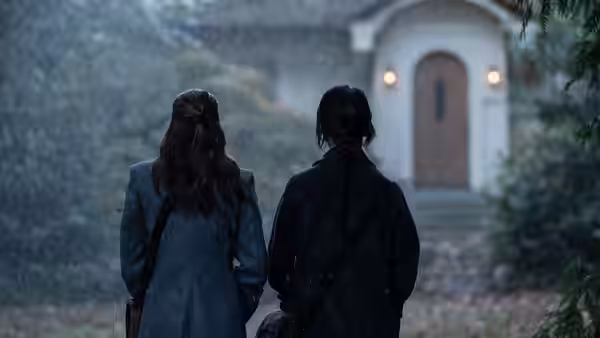 Two women in coats approach a house through falling rain