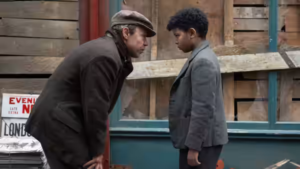 Man in cap speaking to George (Elliott Heffernan) outside a building