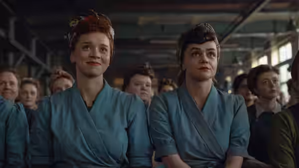 Two women in blue overalls seated among factory workers