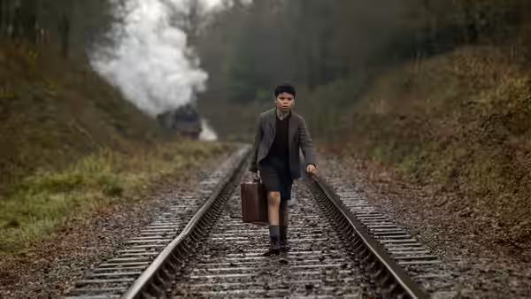 George (Elliott Heffernan) walking along railway tracks carrying a suitcase