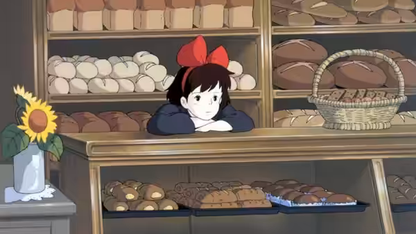 Kiki (Minami Takayama) resting her arms on a bakery counter with bread shelves behind her