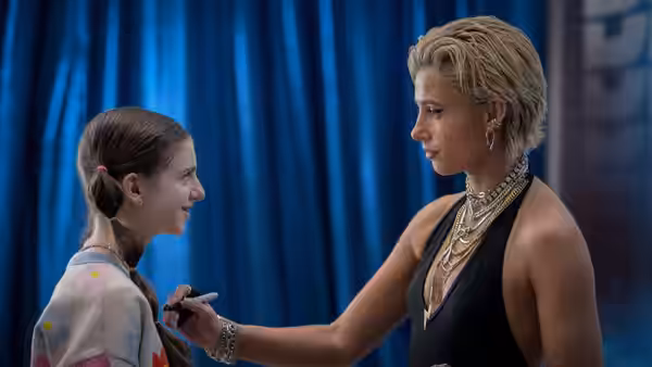 Young girl facing Skye Riley (Naomi Scott) by a blue curtain