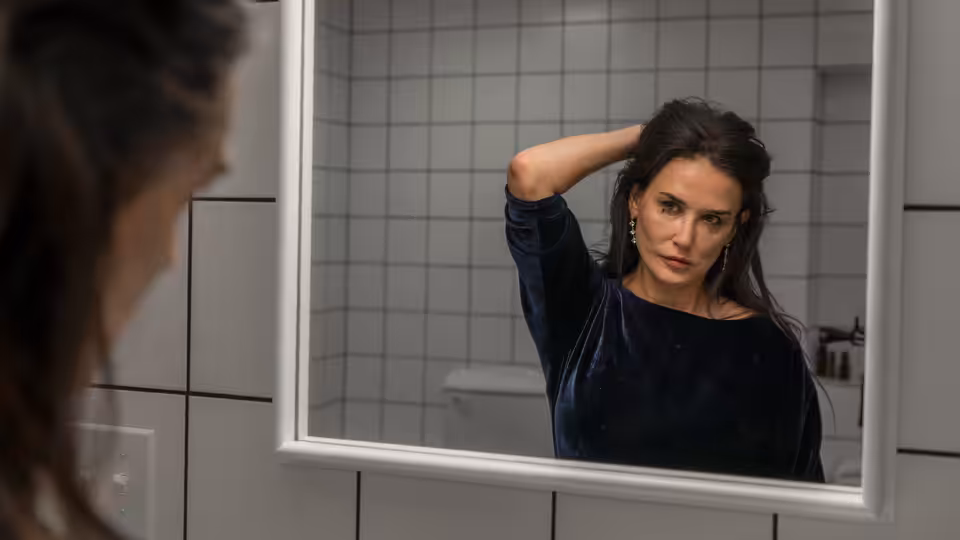Elisabeth (Demi Moore) adjusting her hair while looking in a bathroom mirror