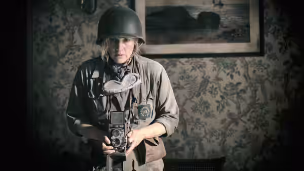 Lee Miller (Kate Winslet) wearing helmet and holding camera in patterned room