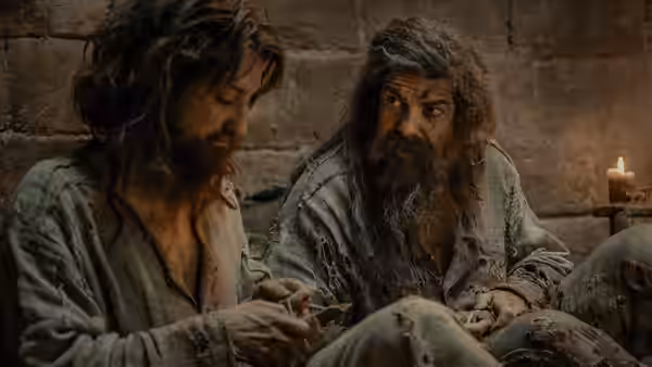 Two disheveled bearded men sit inside a dimly lit stone prison cell