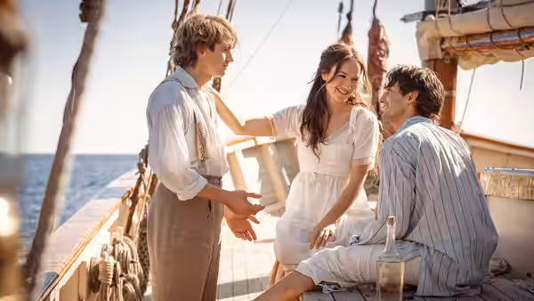 A young woman smiles between two men on the deck of a sailing ship