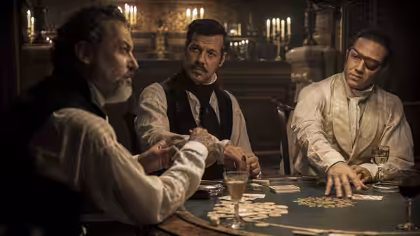 Four men play cards around a table in a candlelit ornate room