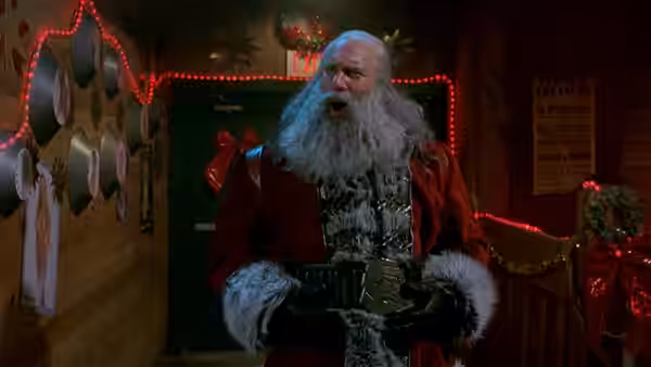 Santa (Bill Goldberg) stands indoors holding two mugs in a decorated room