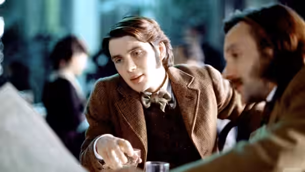 Sir Felix Carbury (Matthew Macfadyen) leans across a table in a crowded room