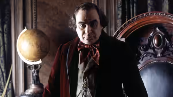 Augustus Melmotte (David Suchet) stands beside a globe and carved chair