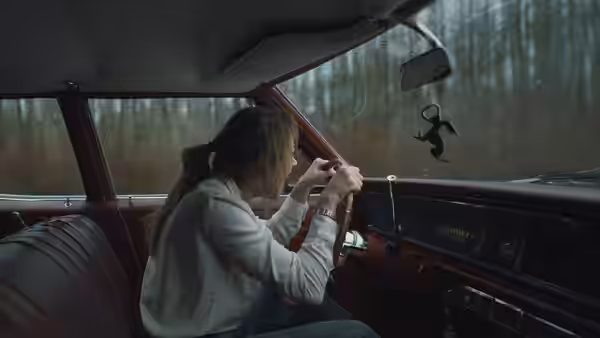 Agent Lee Harker grips the wheel inside a vintage car in woods