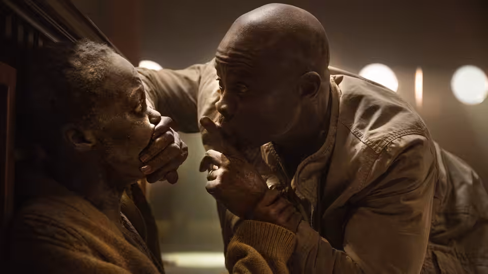 Henri (Djimon Hounsou) covers Samira's mouth as they hide indoors