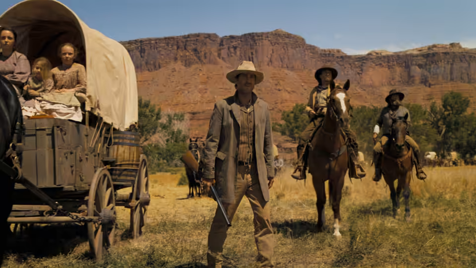 A man with a rifle stands before a covered wagon in the desert while riders approach