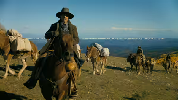 A horseman leads a pack train across a grassy hillside under a clear blue sky