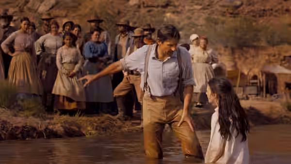 A man gestures to a young girl in a stream as a crowd watches from the shore