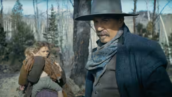 A man in a wide hat stands near a woman holding a child in the woods