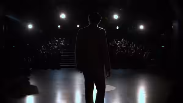 A man in a suit stands alone on a dark stage facing a seated audience