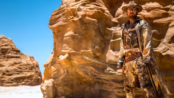 A man in hat and cape holds a sword among desert rock formations