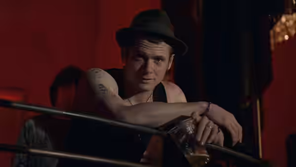 Blake (Jack O'Connell) leaning on a railing holding a drink