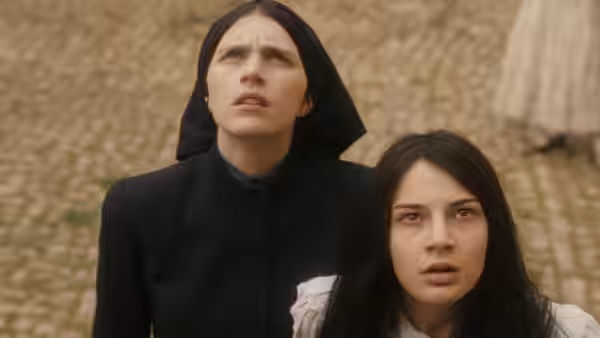 a nun in black habit stands next to a young woman on a cobblestone street