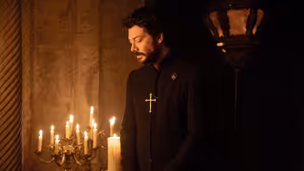 A priest in black clerical attire stands in a candlelit stone room with a cross necklace