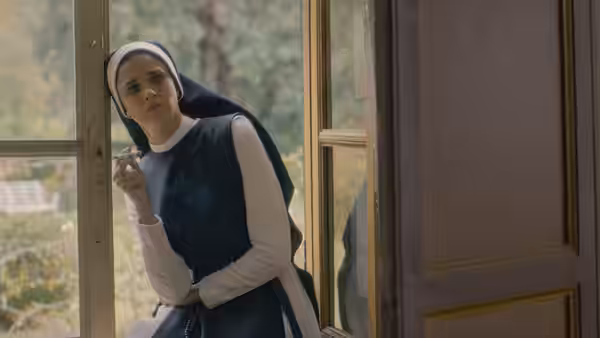 A nun in a habit leans against a window frame while holding a cigarette