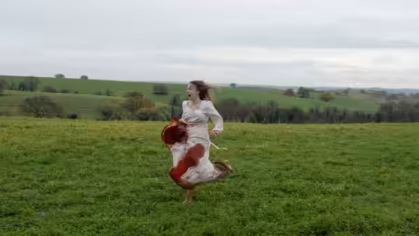 A barefoot woman in a blood-stained white dress runs across a green hillside field