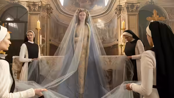 A woman in a blue gown and long veil stands in a church as nuns adjust her train