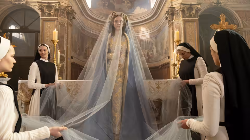 A woman in a blue gown and long veil stands in a church as nuns adjust her train