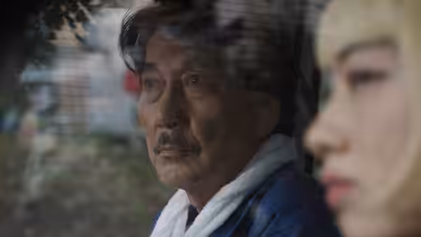 Hirayama (Koji Yakusho) stands outdoors beside a blonde woman looking to the side